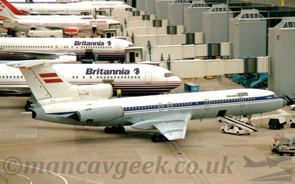 Side view of a 3 engined jet airliner parked by a terminal, facing to the right.
The plane has a white upper fuselage, with grey on the lower section, and a blue stripe running along the body, covering the passenger cabin windows.
There are black "LatAvio" titles on the upper forward fuselage, with smaller white text "Tu154B-2" on the blue stripe just aft of the cockpit.
The black registration "YL-LAC" is on the centre engine intake, just in front of the tail, which itself has a large Latvian flag (2 thick red horizontal stripes separated by a thin white stripe) in the middle.
The other engines, one on each side mounted on the rear fuselage, are grey.
A baggage compartment hatch is open in the lower fuselage, between the forward- and mid-cabin doors, with a mobile baggage-loading belt raised  towards it.
Grey concrete apron fills the foreground, with a large terminal building on the right of the frame, and several mostly white airliners parked nose in, with white airbridges attached to the far sides.