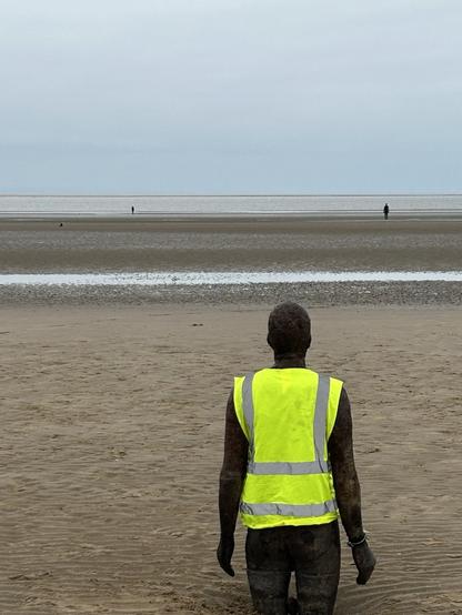 Photo of iron man statues standing in the sand looking out to sea. The one nearest the camera is wearing a reflective jacket.