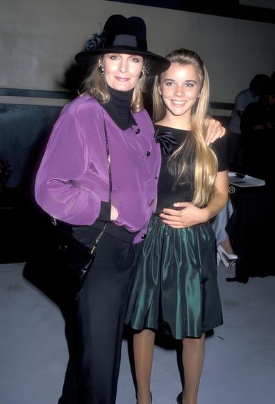 A 1988 photo of Deidre Hall and Christie Clark, best known for their roles as Marlena Evans and Carrie Brady Reed on Days of Our Lives, taken at the Starlight Children’s Foundation Benefit. Deidre wears a purple satiny jacket with black trim over black pants, accessorized with a black hat. Her shoulder-length light brown hair is styled in soft waves. Christie wears a black cocktail dress with a bow detail, and her long blonde hair is styled with an alice band. Both are smiling warmly at the camera. The background features a black and beige wall, suggesting an indoor event setting. The image captures a moment of warmth, legacy, and 1980s fashion flair
