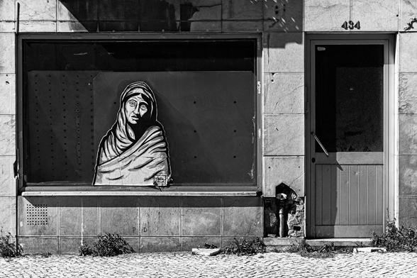 Fotografia a preto-e-branco da montra de uma loja devoluta. No centro da montra pode ver-se um paste-up do artista Tinta Crua, representando uma mulher que enverga um hijab. O olhar vazio e a expressão desconsolada do seu rosto evidenciam um intenso sofrimento.