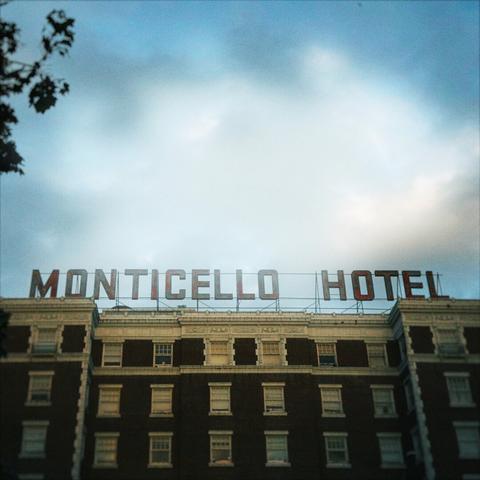 Giant metal lettering sits supported above a large brick building that reads, "MONTICELLO HOTEL," under a hazy sky.