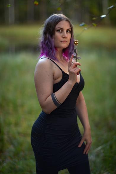A finnish woman wearing a tight black dress posing in front of a pond in an autumn forest looking at camera