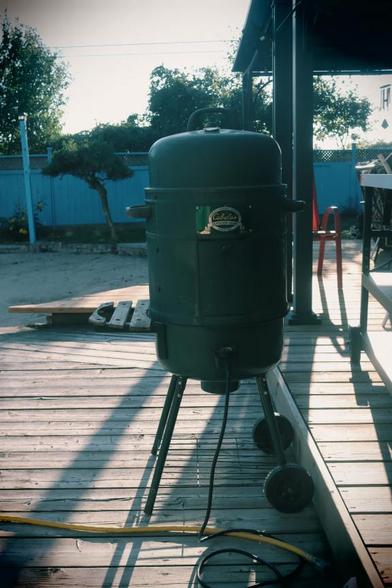 A vertical electric water smoker rests on a backyard wood deck. In the far background is a wooden fence, trees rise up behind the fence.