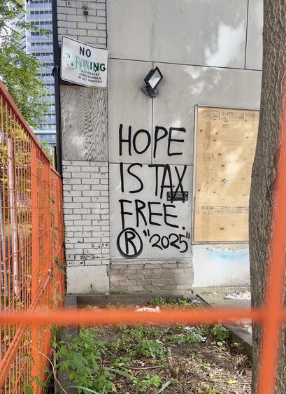 Hope is tax free (R) 2025 black spray paint on boarded up building behind an orange construction fence
