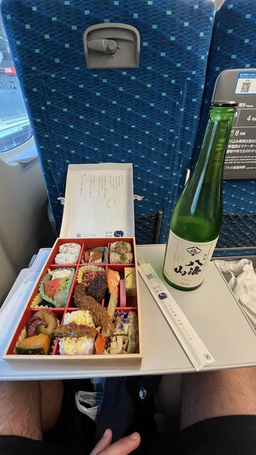 Photo of a bento box and sake bottle from a train seat. The bento box is square with 9 smaller squares within, each with a different morsel of food.