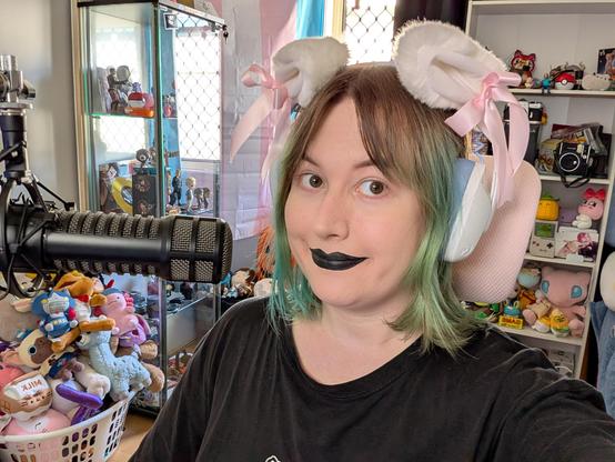 Revy smiling and wearing black lipstick and cat ear headphones 
Many plushies in background too