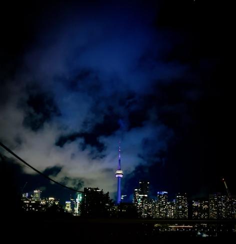 Skyscrapers and CN Tower lit up at night, top 2/3 of the photo is a dark blue sky with one large cloud silhouetted by city lights.