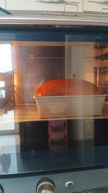 Inside view of an oven with warm, golden light. A silver bread tin in which white bread is being baked. This is towards the end of baking time - the loaf has risen evenly and beautifully, acquiring a deep golden brown colour during the process.