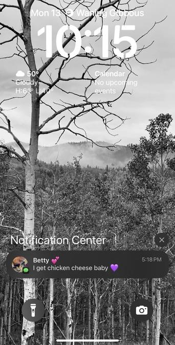 A smartphone screen displaying a black-and-white image of trees and mountains, with a notification from a contact named Betty about "chicken cheese." The time is 10:15, and the weather is cloudy at 50°F.