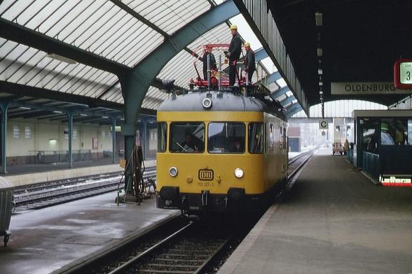Ein Bild aus der Halle eines Bahnhofs, aufgenommen an einem bedeckten Tag, zeigt ein besonderes, gelbes Schienenfahrzeug auf einem Gleis.
​Das Fahrzeug, eine kleine, gelbe Lokomotive mit einer hohen Kabine, steht rechts auf dem Gleis, das dem Bahnsteig am nächsten liegt. Auf dem Dach der Lokomotive stehen drei Männer in Arbeitskleidung und Schutzhelmen, die an roten Geräten arbeiten. Vorne trägt die Lokomotive die Aufschrift "DB" und leuchtende Scheinwerfer.
​Das Gleis befindet sich rechts neben einem breiten Bahnsteig. Über dem Gleis und dem Bahnsteig erstreckt sich eine große, gewölbte Dachkonstruktion aus Glas und dunkelgrünen Metallstreben.
​Rechts in der Ferne ist ein Schild mit der Aufschrift "OLDENBURG" zu erkennen. Links vom Fahrzeug ist ein weiterer Bahnsteig unter der Halle zu sehen, der leer ist. Die Szene vermittelt den Eindruck von Wartungsarbeiten, die an einem Tag ohne Zugverkehr durchgeführt werden.