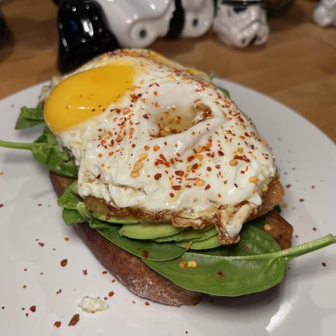 Na zdjęciu tost z ciemnego pieczywa z warstwami świeżego szpinaku, plasterkami awokado, wędzonym łososiem i sadzonym jajkiem posypanym płatkami chili.