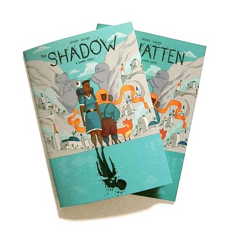 A photo of the printed English and German editions of my comic „The Shadow“, showing the two main characters in front of a city in a valley. They're standing (on the shore?) of a lake. There's a shadowy creature instead of the reflection of one of the main characters in the water.