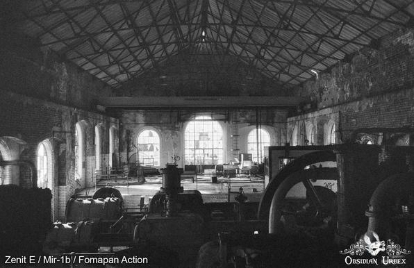 Black and white photo of an abandoned industrial building in with large arched windows, machinery, and a high metal truss roof