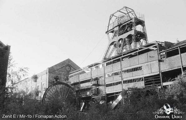 Black-and-white photography of an abandoned industrial site, featuring a towering metal mining headframe and overgrown vegetation in the foreground.