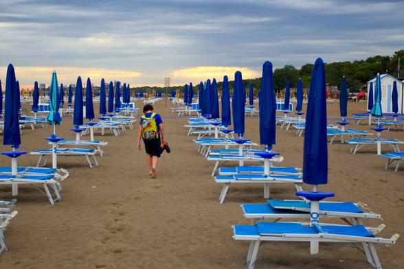 A young person walks barefoot down a wide, empty beach lined with neat rows of closed blue umbrellas and matching sun loungers. They wear a bright green backpack and carry their shoes in one hand, leaving faint footprints in the sand. The sky is cloudy and muted, with a hint of golden light near the horizon, suggesting the day is ending
