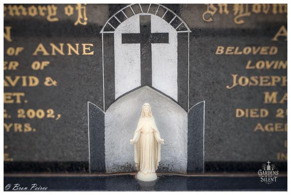 A close up photograph of a statue of the Virgin Mary (Madonna) set into a carved niche on a dark, polished headstone.   The small, white or ivory colored figure stands at the base of the niche, which is outlined in black and white stone to form a pointed, Gothic archway.  Above the figure, a simple cross is carved into the stone. The surrounding surface of the headstone is a dark, glossy material with golden, engraved text visible on either side and blurring into the background.  The bottom left corner is signed "© Bron Peirce."