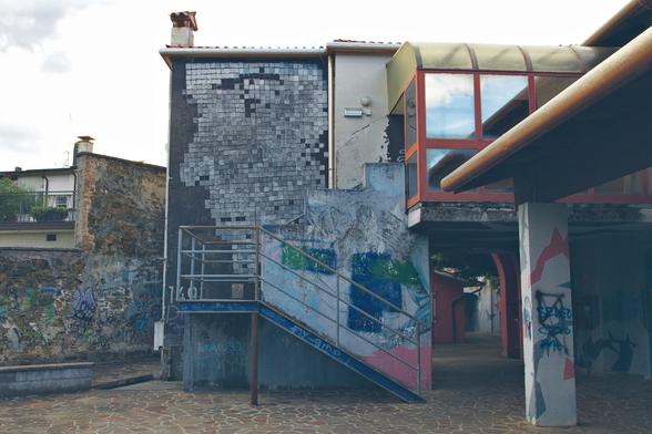 A weathered building with cracked walls and faded graffiti stands under a cloudy sky. A metal staircase leads up to a small bridge-like corridor with orange-framed glass panels. The side of the building features a large mosaic or mural in grayscale tiles, partly deteriorated.