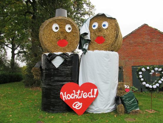 Hochzeitspaar aus großen Strohballen an einer Straße. Rechts daneben eine kleine Figur, die an der Braut lehnt.