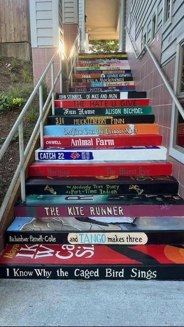 The books depicted on the staircase include:

1984 by George Orwell

To Kill a Mockingbird by Harper Lee

Fahrenheit 451 by Ray Bradbury

Of Mice and Men by John Steinbeck

The Hate U Give by Angie Thomas

Fun Home by Alison Bechdel

The Adventures of Huckleberry Finn by Mark Twain

One Flew Over the Cuckoo's Nest by Ken Kesey

Animal Farm by George Orwell

Catch-22 by Joseph Heller

The Bluest Eye by Toni Morrison

The Absolutely True Diary of a Part-Time Indian by Sherman Alexie

The Kite Runner by Khaled Hosseini

And Tango Makes Three by Justin Richardson, Peter Parnell, and Henry Cole

I Know Why the Caged Bird Sings by Maya Angelou