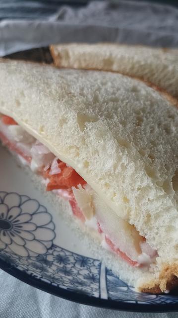A close up of the cut edge of a white bread sandwich showing the crumb and fillings (potato, tomato etc) kept on a white plate with blue floral design.