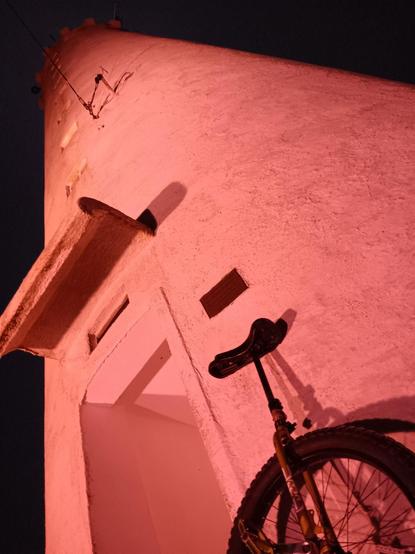 A low angle view of a lighthouse illuminated in pink light, looking upwards, with a unicycle leaning against its base. Taken in the very early morning, so it is dark outside.