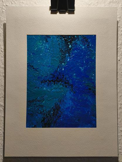 Fotografie eines abstrakten Acryl Gemäldes mit einer dominierenden Farbpalette aus verschiedenen Blautönen, teilweise mit metallischem Glanz. In der Mitte des rechteckigen Kunstwerks treffen tiefblaue und türkisfarbene Flächen in unregelmäßigen, teils scharf abgegrenzten, teils weich verlaufenden Übergängen aufeinander. Dunklere Bereiche, fast schwarz, strukturieren die Komposition diagonal von der oberen rechten zur unteren linken Ecke und erzeugen eine dynamische Bewegung. Die Oberfläche ist stark strukturiert und reliefartig, mit sichtbarer pastoser Farbauftragung, die das Licht unterschiedlich reflektiert. Das Bild ist mit glänzendem Acryl Finish endbehandelt. Das Kunstwerk ist von einem breiten Passepartout umgeben und hängt an einer hellen Wand.