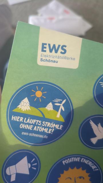 Das Foto zeigt einen grünen Flyer mit Stickern. Oben steht: EWS ElektrizitätsWerke Schönau. In der Mitte ist ein blauer, runder Aufkleber   mit zwei Hügeln, zwei Bäumen, einer gelben Sonne und einem Windrad. Darunter steht: Hier läufts Strömle ohne Atömle!