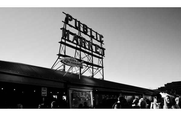 Black and white photo of a bustling public market under a clear sky. Prominent "Public Market" sign above, people walk below, evoking a lively atmosphere.