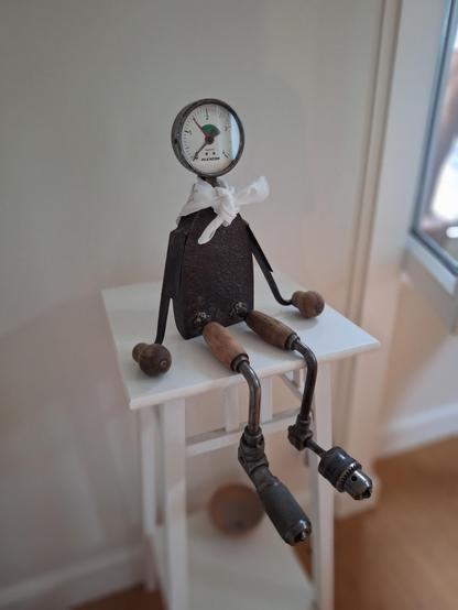 Personnage assis sur un tabouret blanc, ses pieds pendent dans le vide. Tronc en fer à repasser ancien, bras en lames et manche de racloirs, jambes et pieds en morceaux de vilebrequins, tête en cadran de détendeur, petit foulard blanc autour du cou
