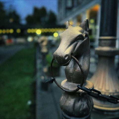 A small, metal horse's head tops a chain fence post in front of a hotel building, with green grass and a twilight sky in the distance.