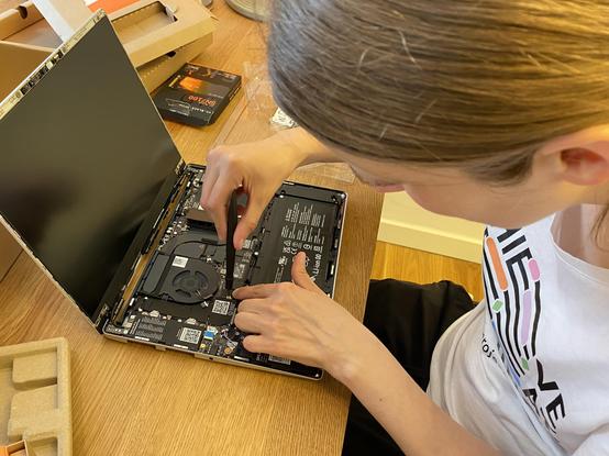 A person is working on a laptop, with the cover removed, examining the internal components. Tools are visible, and there are boxes and packaging materials nearby. The setting appears to be a home or office with a wooden table.