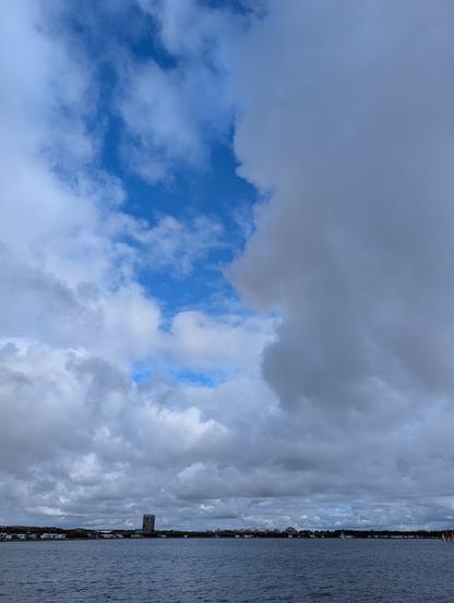 Das Bild zeigt eine Landschaftsaufnahme mit einem großen Wasserbereich im Vordergrund und einem Horizont mit Land im Hintergrund. Der Himmel nimmt den größten Teil des Bildes ein und ist von Wolken bedeckt, wobei zwischen den Wolkenabschnitten blaues Licht zu sehen ist. Die Wolken erscheinen dunkel und dichter in den Randbereichen und heller in der Mitte. Am Horizont ist eine Silhouette einer Stadt oder einer Ansammlung von Gebäuden mit einer einzelnen hohen Struktur erkennbar. Das Wasser ist relativ ruhig und spiegelt das dunkle Licht des Himmels wider.
