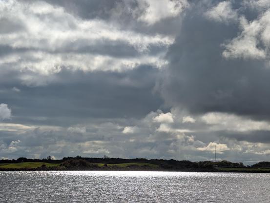 Ein horizontales Bild zeigt eine Landschaft mit einem großen Gewässer im Vordergrund. Das Wasser ist glitzernd und reflektiert das Licht. Über dem Wasser befindet sich eine flache, dunkle Landmasse mit Bäumen und Büschen. Der Himmel ist bedeckt von dunklen Wolken mit helleren Flecken, die das Licht durchscheinen lassen. Ein hoher Antennenturm ist auf der Landmasse zu sehen.

Bereitgestellt von @altbot, privat und lokal generiert mit Gemma3:27b