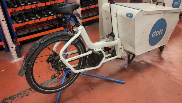 Vélocargo Urban Arrow avec la caisse avant floquée Dott dans un entrepôt. Le vélo est accroché à mon pied d'atelier ce qui soulève la roue arrière.
En arrière plan, des batteries de trottinettes en train de charger