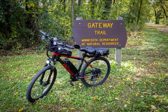 A black and red electric bike in front of a brown wooden sign that says "GATEWAY TRAIL - MINNESOTA DEPARTMENT OF NATURAL RESOURCES" in yellow letters