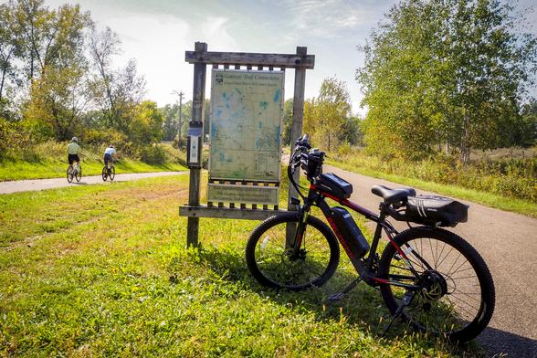 A black and red electric bike in front of a large map of the Gateway Trail.  Two cyclists are on the path in the background.