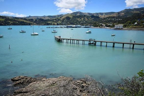 Au premier plan, on aperçoit un petit promontoire rocheux bordant une eau claire, presque immobile. Fait de bois, il s’avance de la rive jusqu’à la mer, soutenu par des pilotis fins.
Plusieurs voiliers blancs sont ancrés dans la baie, immobiles sur l’eau d’un bleu laiteux. À l’arrière-plan, les collines douces et vertes se déploient sous un ciel bleu vif, parsemé de quelques nuages blancs.
De petites maisons blanches et des bâtiments épars ponctuent le rivage, évoquant un village tranquille au bord de l’eau.
L’atmosphère générale est calme, lumineuse, presque méditative — une impression de sérénité absolue, comme un matin d’été dans un port lointain.