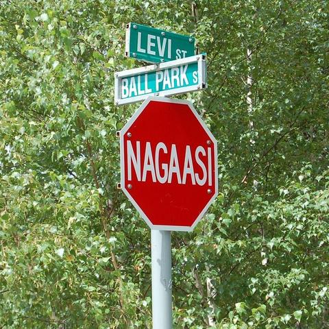 A red octagonal stop sign with the word “NAGAASI” written in white letters, located at the intersection of Levi Street and Ball Park Street. The background is filled with green leafy trees.