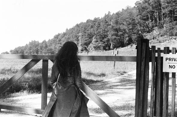 A woman with long hair and a long coat is seen from behind approaching a gate in front of a wooded landscape. A sign reading “Private. No trespassing” can just be seen to the right.