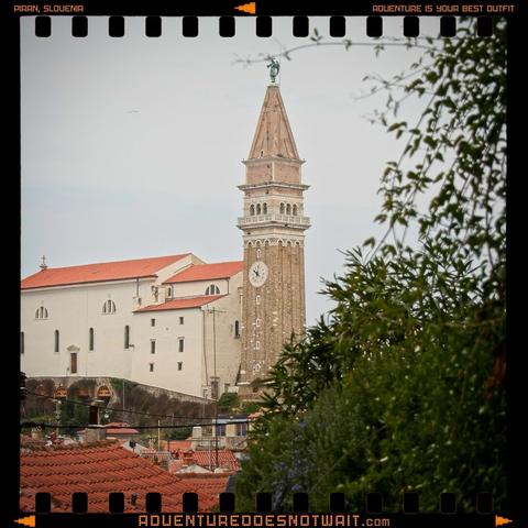 A historic clock tower rises above terracotta rooftops in Piran, Slovenia, framed by lush greenery. The timeless scene evokes the organic textures and earthy tones of sustainable adventure apparel.