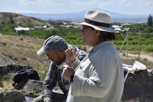 Due archeologi discutono sul sito di scavo in Argištiḫinili, Armenia, con un paesaggio di colline e abitazioni sullo sfondo.