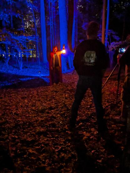 A figure in a long red cloak, holding aloft a small lantern, standing in the woods while two figures on the right side record the scene on a camera.
