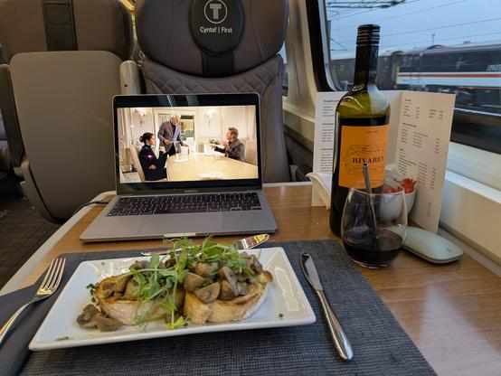 A picture of my table in first class on a Transport for Wales train. A bottle of red wine is on the right next to a broad-base glass with a metal straw in.  A placemat is laid out with cutlery and a plate with garlic mushrooms on toasted bread in front of a Macbook Pro open with Only Murders in the Building playing.