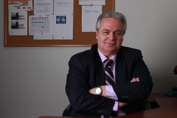 El autor de la columna, Juan Francisco Ortún, vestido con un traje oscuro y una camisa clara, sentado en un escritorio con las manos cruzadas y una expresión seria, frente a un fondo de documentos en un tablero de corcho.