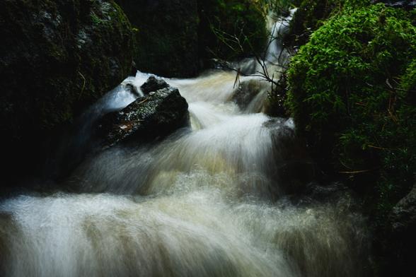 A photo of a very small waterfall in a little forest stream. The waterfall runs down the middle of the image, consisting of several levels with the water zig-zagging down.

The fall is surrounded by stones, whilly or partly covered in green moss.

The photograph is taken with a moderately long exposure time, giving the water a partly silky appearance as it rushes down.