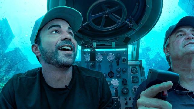 Two men inside a two-person submarine surrounded by murky blue-green water, with the pilot pointing upward toward instrument gauges on the control panel.