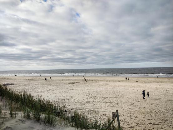 Ein später Herbstnachmittag am Strand, im Hintergrund die Nordsee. Der Himmel ist bewölkt, hier und da scheint ein wenig Blau durch die Wolkendecke. Am Strand sieht man vereinzelt Personen beim Spaziergang.