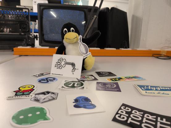 Ein Tux-Plushie sitzt auf einem Tisch. Davor diverse Themeverwandte Sticker, darunter Conversations, I2P, TOR, Qutebrowser, La Quadrature du net. Im Hintergrund ein Röhrenfernseher.