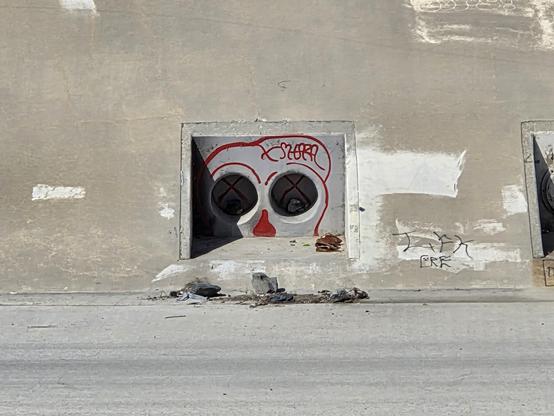 Zoomed in photo on a water outlet inset in a large concrete channel. Two side-by-side water pipes look like eyes and graffiti has been added around them to appear like they belong to a skull.