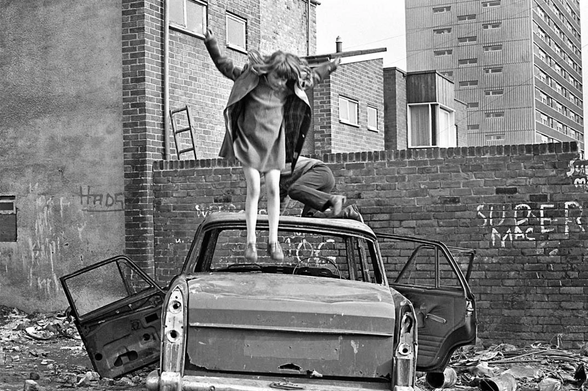 Photographie anglaise en noir et blanc de deux enfants qui sautent sur une voiture abandonnée. En fond, des barres d'immeuble